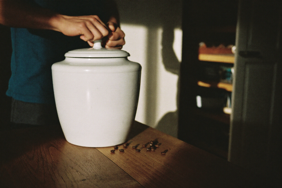 Hands pressing the lid onto an opaque stoneware jar of coffee beans on a kitchen counter, a few loose beans beside it.