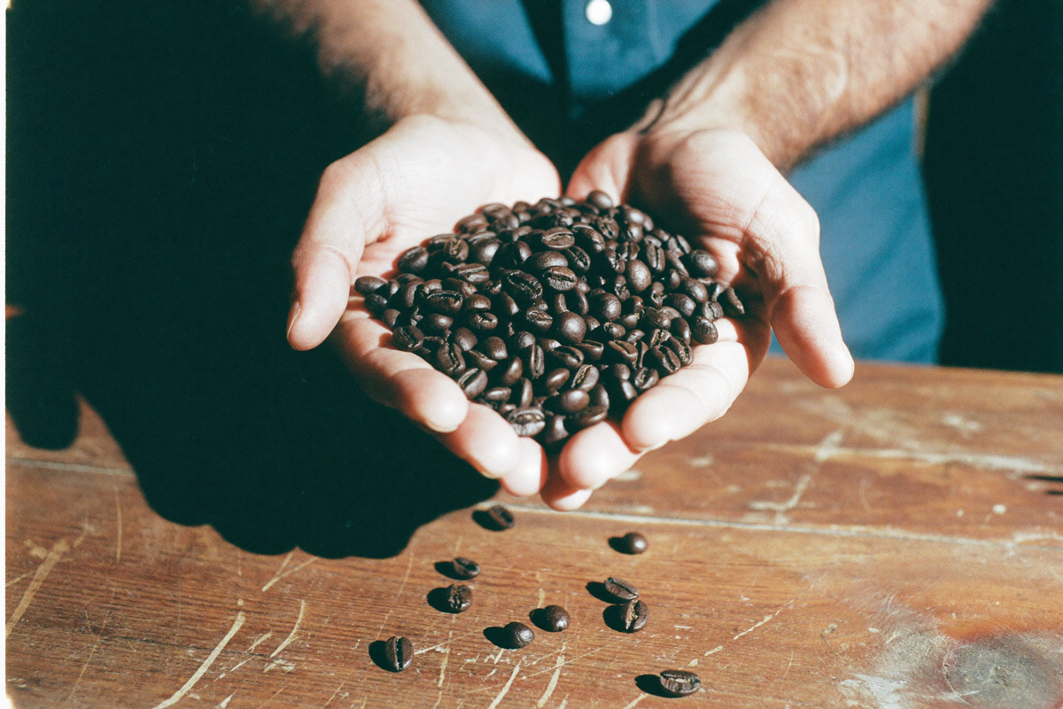 Two cupped hands holding a mound of dark roasted beans over a scarred wooden table, a few beans fallen onto the wood.