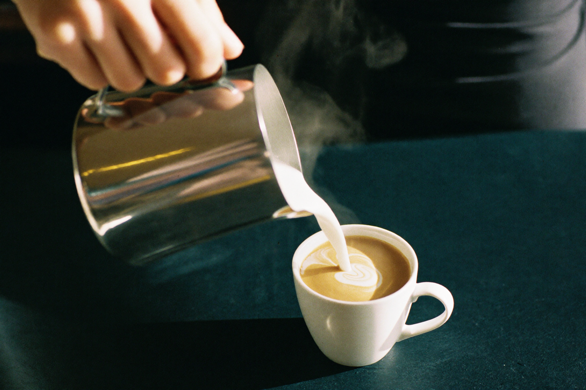 A hand tilting a plain steel jug to pour steamed milk into a white cup, a swirl opening across the dark surface.