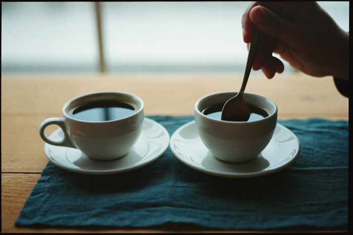 Two identical white cups of coffee side by side on teal linen, a hand lifting a spoon from the right-hand cup.