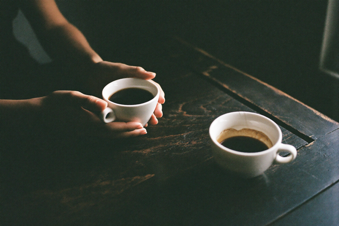 Two small white cups of black coffee on a dark scarred wooden table, a pair of hands resting around one of them.