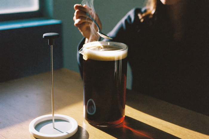 A hand skimming foam from the surface of a glass coffee press, its plunger and lid set aside on the table.
