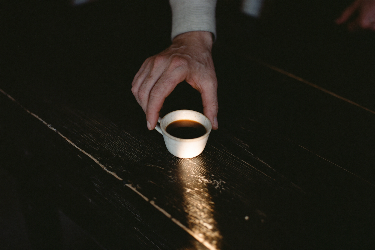 A single small handleless cup of very dark coffee on near-black wood, one narrow shaft of evening light across it.
