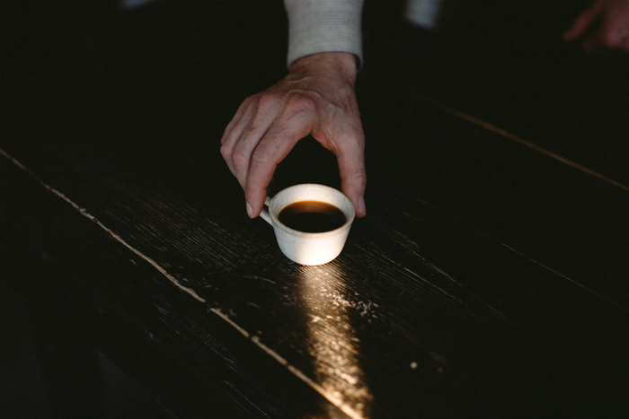 A single small handleless cup of very dark coffee on near-black wood, one narrow shaft of evening light across it.