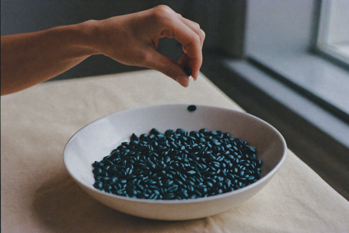 A shallow white bowl of roasted beans on linen, a hand lifting a few out between finger and thumb.