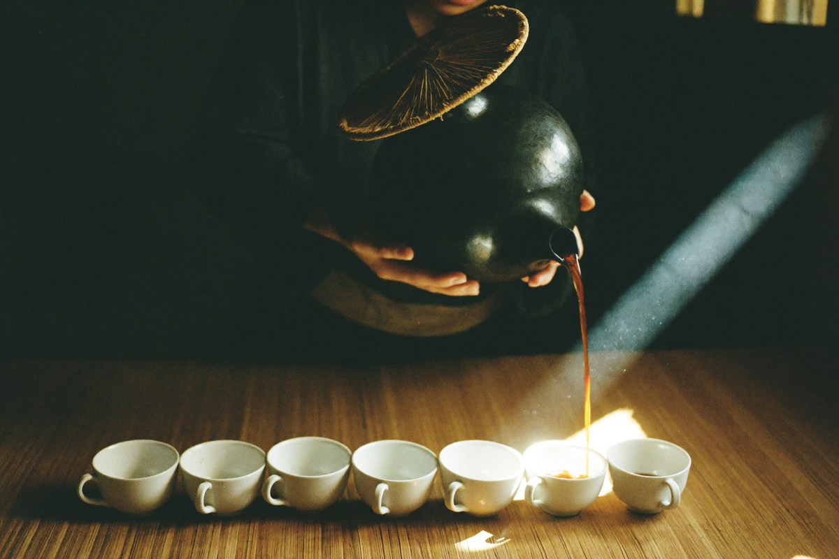 A black clay pot with a straw lid poured from height in a long arc into a row of small white cups.