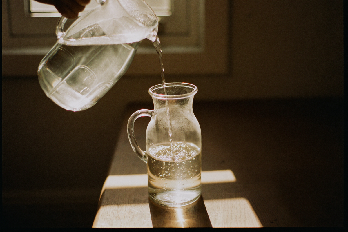 Water poured from a plain glass jug into a glass carafe on a wooden table, lit hard from behind.