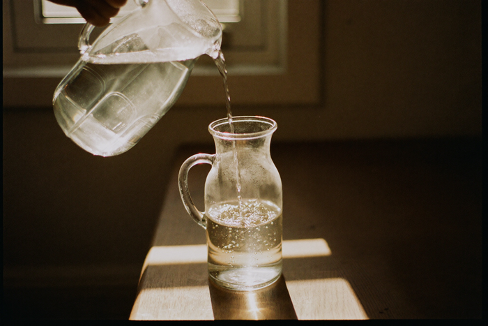 Water poured from a plain glass jug into a glass carafe on a wooden table, lit hard from behind.