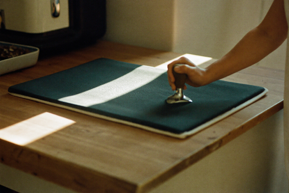 A folded dark rubber mat on a worn wooden counter with a heavy flat-based tamper being set down on it.