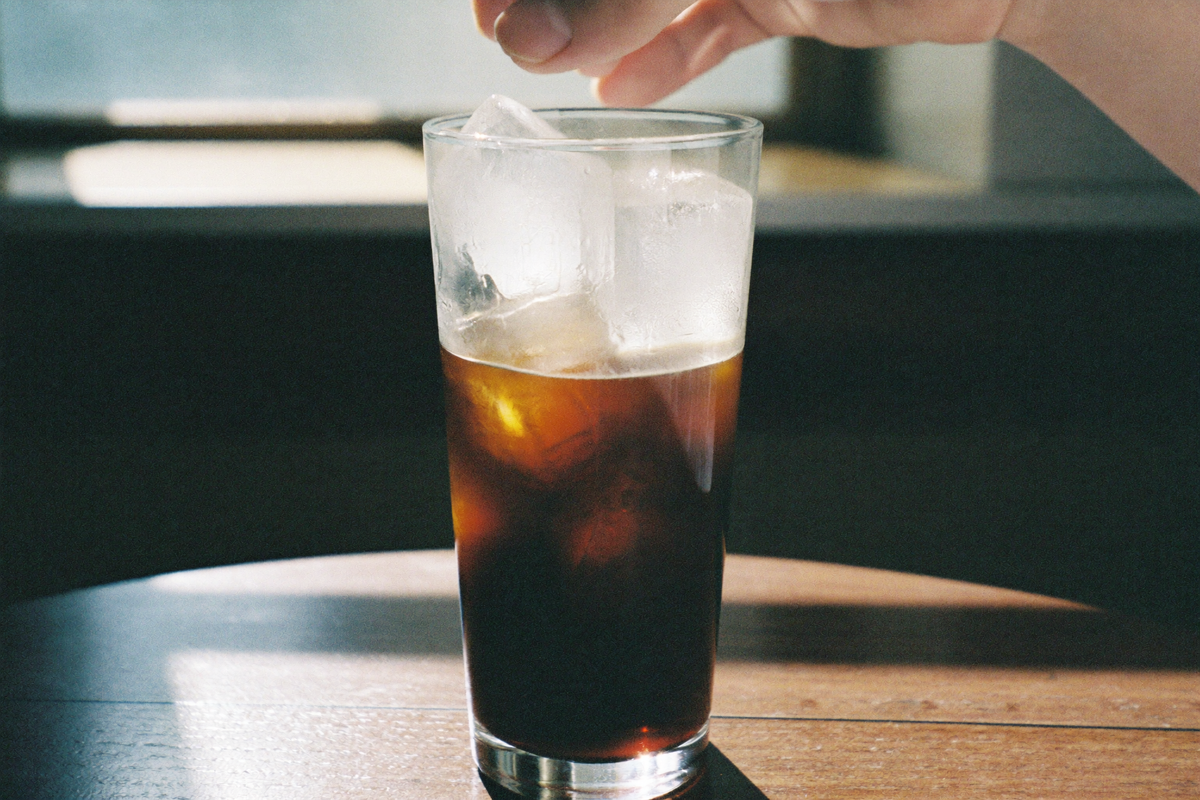 A tall glass of very dark cold coffee over clear ice, condensation running down the outside as a hand lifts it.