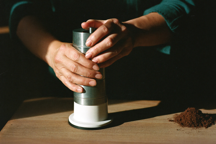 Two hands gripping and turning the body of a cylindrical steel hand grinder, ground coffee heaped on the wood beside it.