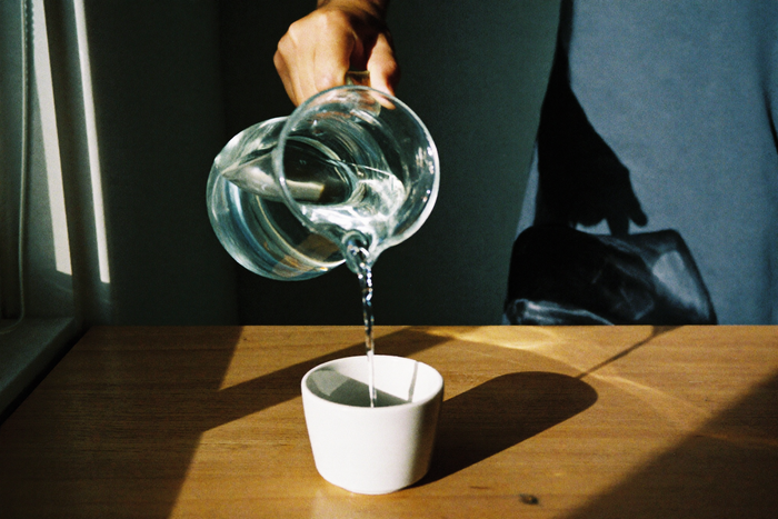 A hand pouring a thin stream of water from a clear glass jug into a white ceramic cup on a wooden counter.