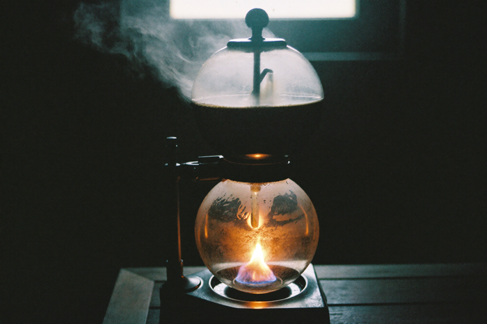 A two-chamber glass vacuum brewer with a small flame beneath, coffee suspended in the upper globe, the room dark.
