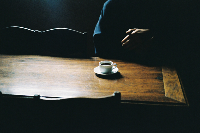 A small white cup of black coffee on a heavy dark refectory table, an older pair of hands folded beside it.