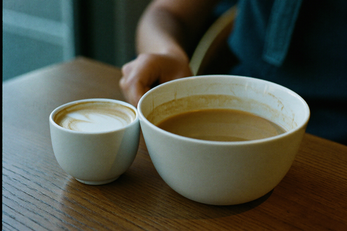 A small thick-walled cappuccino cup standing beside a much larger bowl-like cup, both with a fine foam surface.